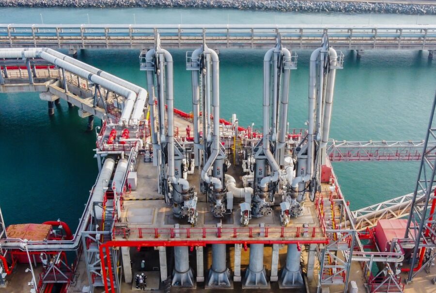 Industrial offshore processing platform with silver pipes and red support frames over calm turquoise water.