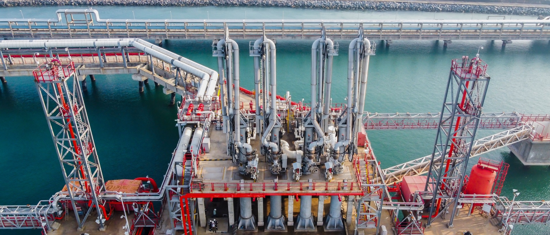 Industrial offshore processing platform with silver pipes and red support frames over calm turquoise water.