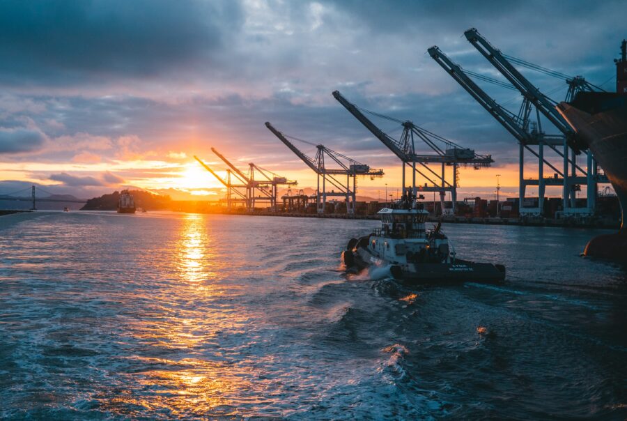 Port cranes silhouetted against a sunset over calm water with a cargo ship nearby, conveying a busy shipping terminal at dusk.