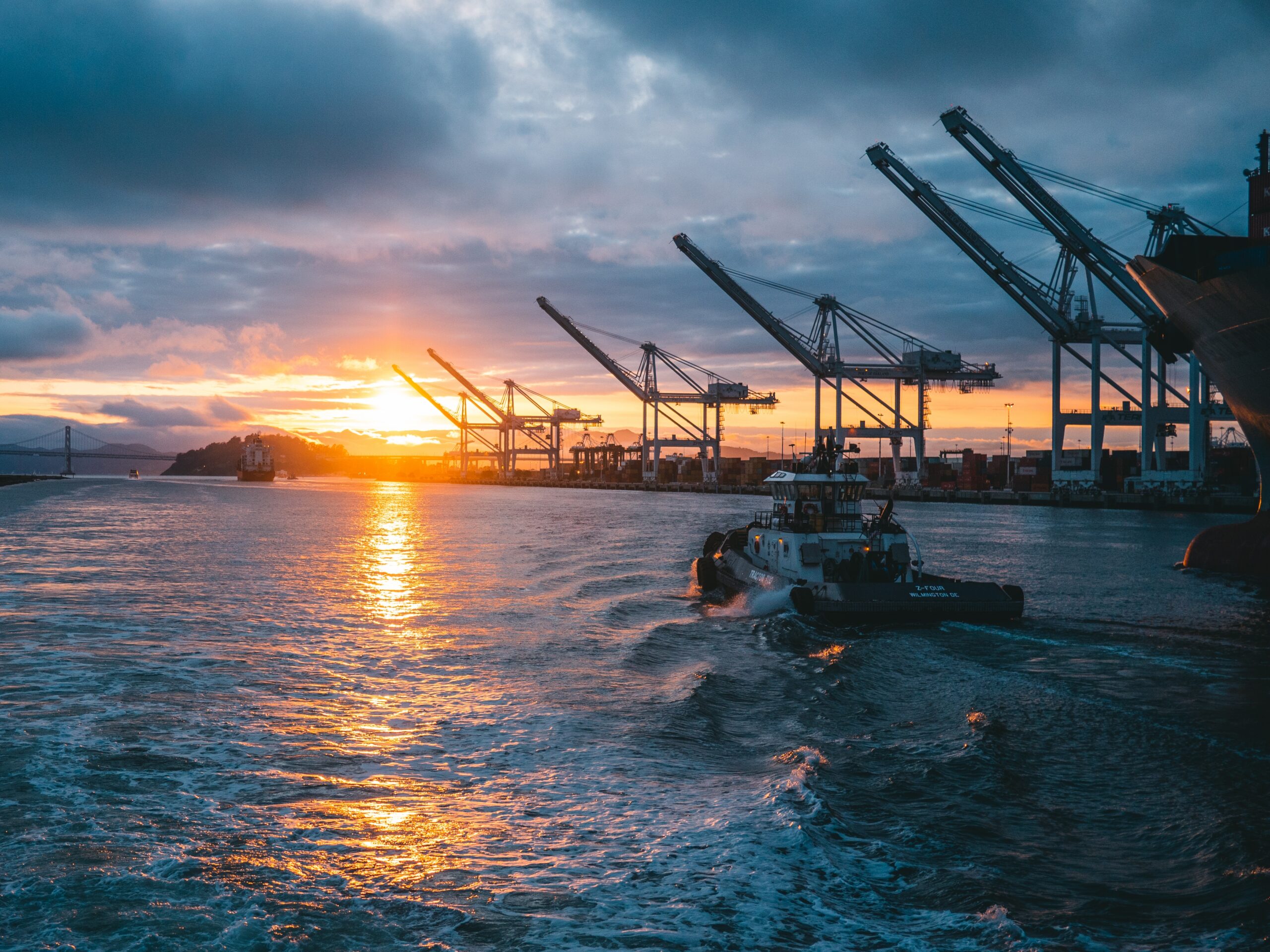 Port cranes silhouetted against a sunset over calm water with a cargo ship nearby, conveying a busy shipping terminal at dusk.